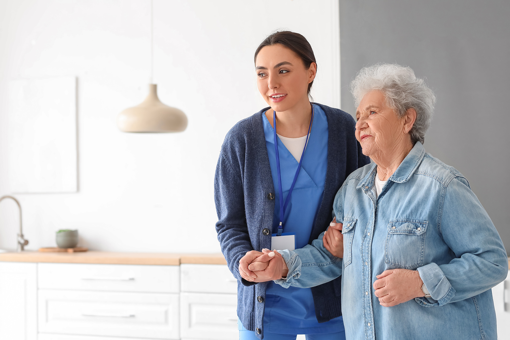 Young caregiver helping senior woman to walk in kitchen